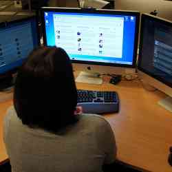 An office worker gazes at an active computer screen, while two other screens are dim.