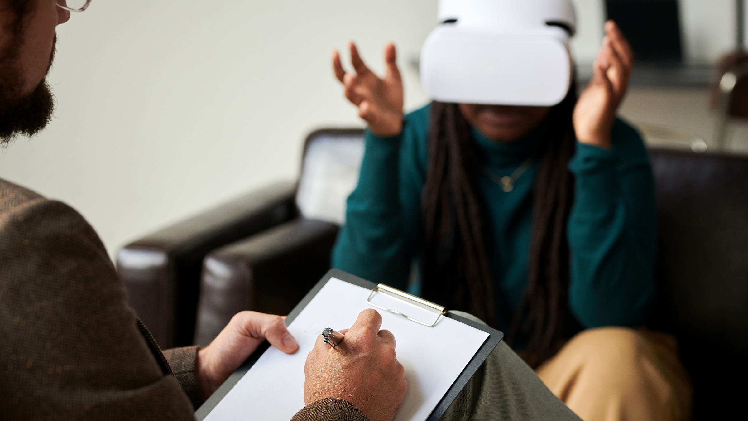 seated person wearing a VR headset gestures in front of a seated man taking notes