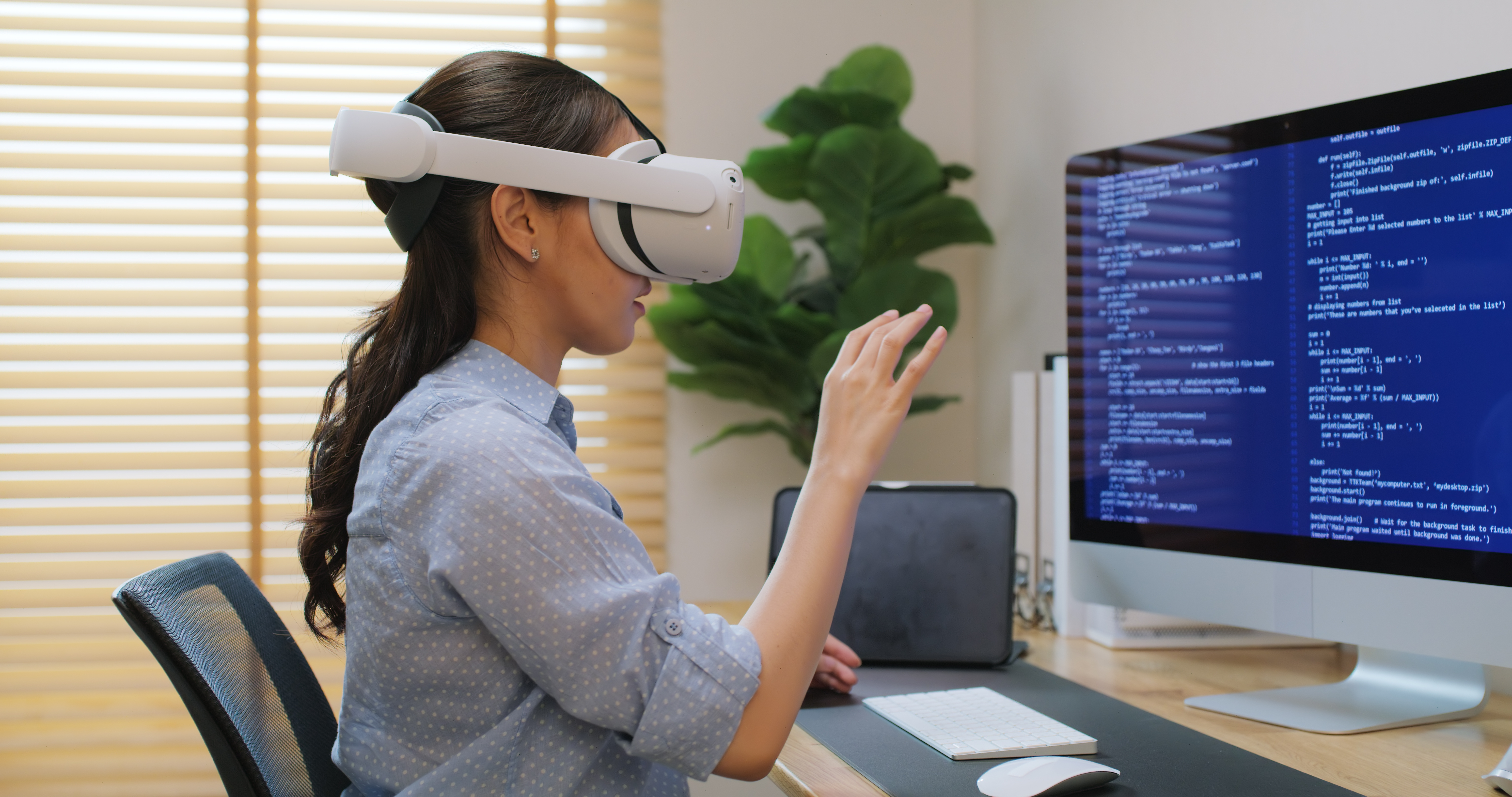 A woman sitting at a desk behind a computer works on coding project while wearing VR goggles.